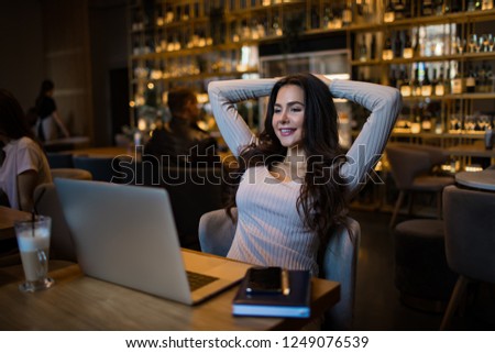 Young charming smiling woman having online video conversation via modern laptop computer while relaxing in restaurant during recreation time. Joyful pretty female watching funny video via notebook 