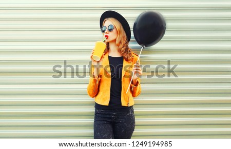 Portrait stylish woman drinking fruit juice holding black air balloon in round hat, yellow jacket on metal wall background