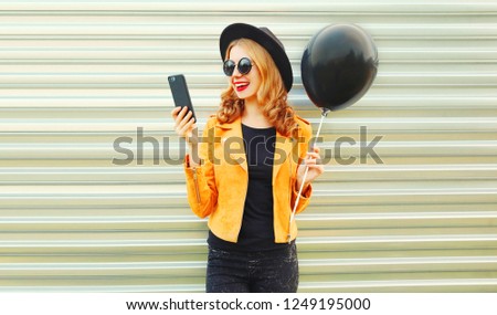 Portrait happy pretty smiling woman with phone holding black helium air balloon in round hat, yellow jacket on metal wall background