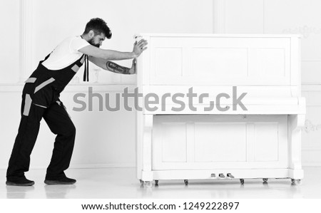 Man with beard and mustache, worker in overalls pushes piano, white background. Courier delivers furniture in case of move out, relocation. Courier service concept. Loader moves piano instrument.
