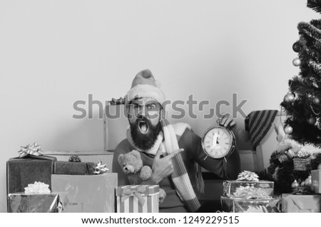 Santa Claus waits for New Year near decorated tree on pink background. Man with beard and shocked face celebrates Christmas. Winter holiday and countdown concept. Santa points at old alarm clock