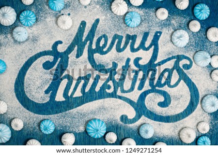Merry Christmas sigh made out of sugar powder surrounded by small blue and white meringue on blue background, flat lay