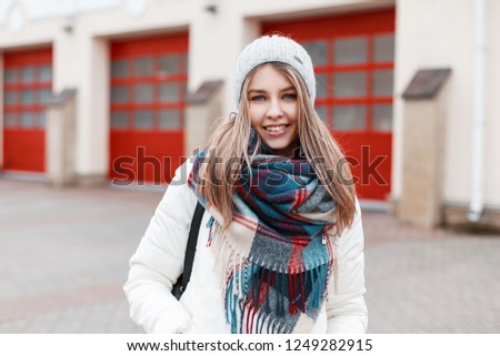 Portrait of a cheerful positive young woman with a sweet smile with a warm stylish checkered scarf and white fashionable jacket against the background of a building with red doors. The girl smiles. 