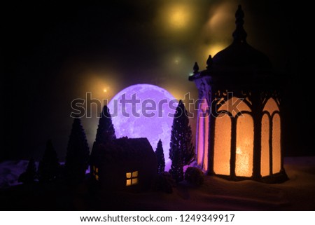 Christmas lantern on snow with miniature house, fir tree and moon at night. Festive dark background. New Year's still-life postcard. Lamp in snow with glowing candle. Holiday concept. Artwork