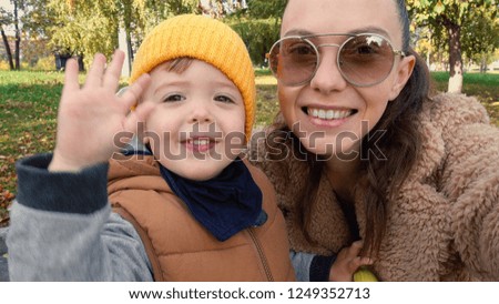 Stylish young woman in sunglasses taking selfie with little son waving with hand at camera in park