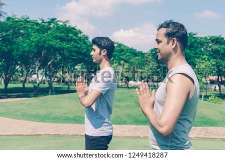 Two serene Indian men meditating and keeping hands together in park with green trees and blue sky in background. Outdoor meditation and yoga concept. Side view.