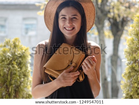 A beautiful smiling brunette poses for a photo dressed in a stylish black dress, a hat and a bag in her hands which she shows in the camera