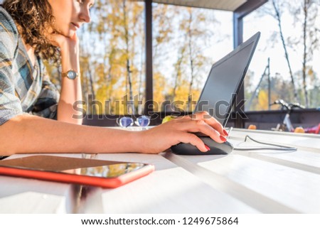 Young woman sitting on outdoor terrace and using laptop. Right hand clicking computer mouse. Sunny day with natural light. Yellow autumn trees on background. Freelance and home office concept. Royalty-Free Stock Photo #1249675864