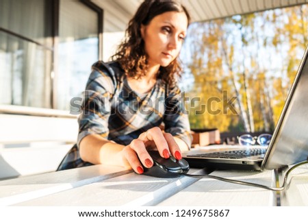 Young woman sitting on outdoor terrace and using laptop. Right hand clicking computer mouse. Sunny day with natural light. Yellow autumn trees on background. Freelance and home office concept. Royalty-Free Stock Photo #1249675867