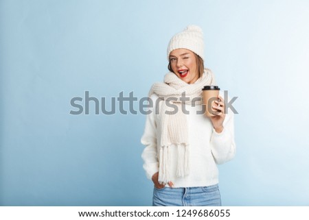 Happy young woman wearing sweater and hat isolated over blue background, holding takeaway coffee