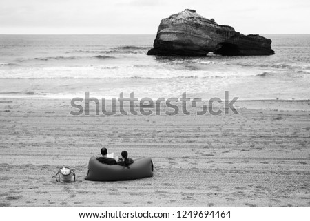 Couple (back view; unrecognizable) relaxing at Biarritz beach (France) in cloudy spring day. Man and woman sitting on air sofa, reading, texting on mobile phone. Relaxation concept. Black white photo.