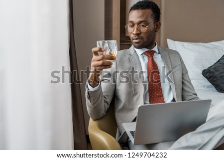 handsome african american businessman with laptop looking at glass of cognac in hotel