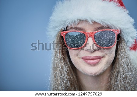 Closeup of frozen girl with snow on face wearing Santa hat and sunglasses, looking at camera smiling
