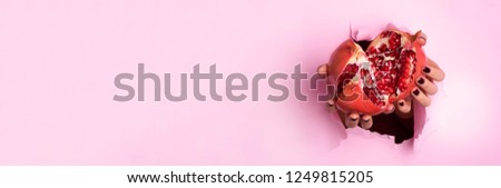 Woman hands holding ripe pomegranate fruit through torn pink paper background. Fresh fruit juice. Vegan, vegetarian concept. Banner with copy space.