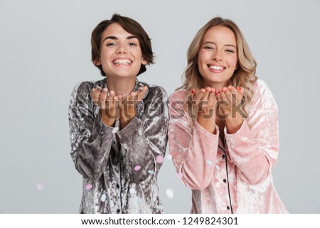 Two lovely girls wearing pajamas isolated over gray background, having fun, playing with confetti