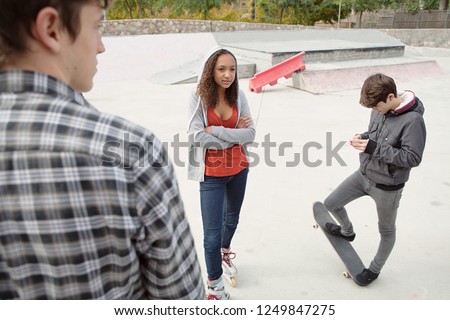 Group of diverse beautiful trendy teenagers in sports ground skating park with skateboard relaxing using technology, outdoors. College students in grunge urban lifestyle, recreation leisure.