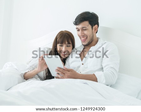 Happy young sweet couple using digital tablet while lying in bed at home. Young happy couple relaxing in bed and connecting with a touch screen tablet, enjoying together.