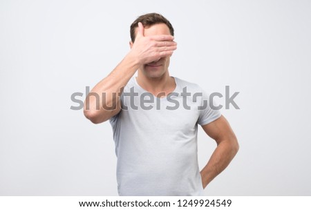 Unrecognizable man with covering eyes while feeling ashamed, standing against blank studio wall background