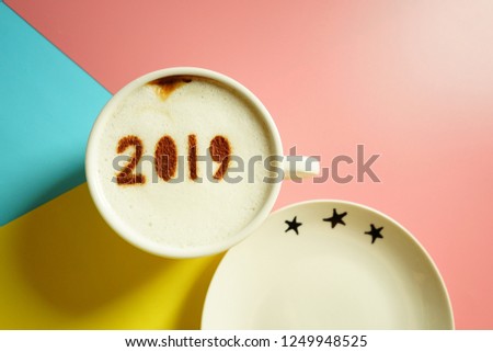 White cup of coffee with the number 2019 on frothy surface over pink yellow and blue colored paper background with three stars saucer. Holidays food art concept for active days in New Year 2019.