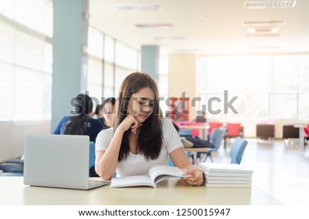 Portrait of young asian female student college is reading book while sitting on table with laptop and stack of textbooks with blurred background of people in library. Education and learning concepts.