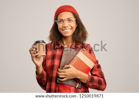 A young smiling woman student in a red plaid shirt and a red hat holds folders with notebooks and documents and a Cup of coffee. Isolated Studio portrait on white background. 