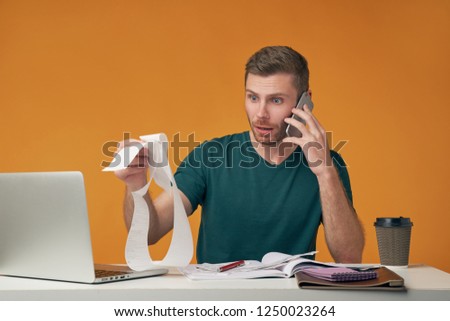 Frustrated young male sitting at table designing new project holding mobile phone in hand, talking to partners. The man considers the cost, surprised by the pricing in the store.
