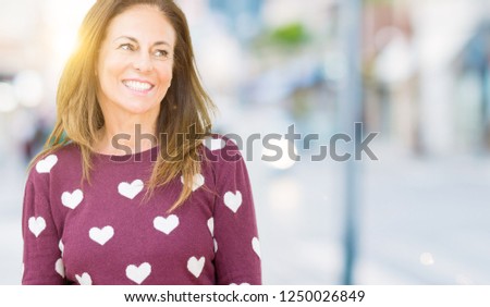 Beautiful middle age woman wearing hearts sweater over isolated background looking away to side with smile on face, natural expression. Laughing confident.