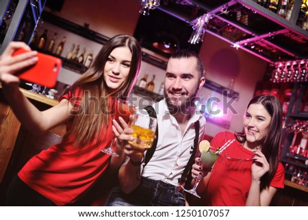 three friends-a young guy and two cute girls holding alcoholic cocktails make a selfie on the background of a bar or a nightclub
