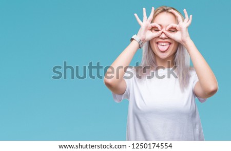 Young blonde woman over isolated background doing ok gesture like binoculars sticking tongue out, eyes looking through fingers. Crazy expression.