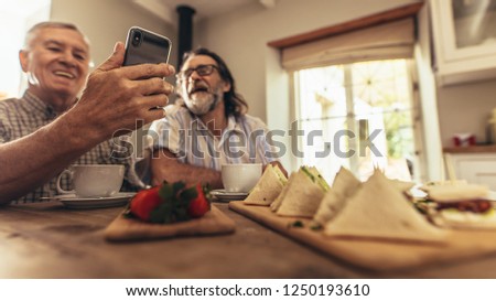Senior men making a video chat with a friends on smartphone. Focus on elderly male hands holding mobile phone while sitting at table indoors.