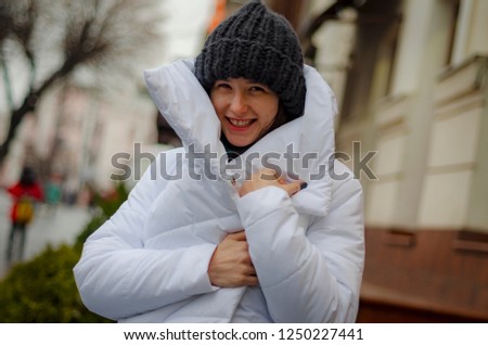beautiful brunette in warm white coat or down jacket on a cold, gloomy street poses with a smile for a photo, an attractive woman in a stylish woolen coat in the winter on the street with a smile