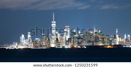 Buildings are illuminated at night looking across the upper bay and Hudson River to the borough of Manhattan