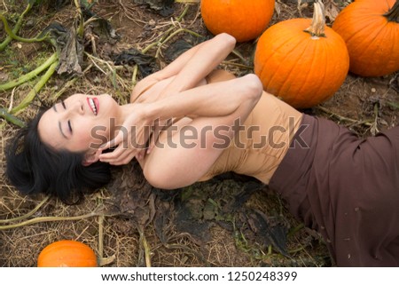 Smiling, half length adult woman dancer dressed in earth tones, lying stretched out among pumpkins in a farm field, in Ellington, Connecticut.