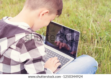 Portrait of smiling well-dressed boy sitting on grass with computer and typing something. Place for text