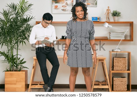 Portrait of African smiling beautiful businesswoman wearing stylish dress standing at office with businessman making notes in his notebook behind her