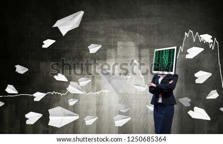 Business woman in suit with monitor instead of head keeping arms crossed while standing against flying paper planes and analytical charts drawn on dark wall on background.