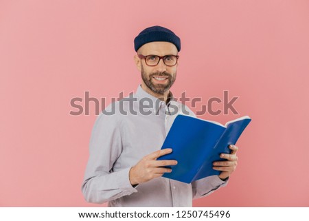 Cheerful unshaven clever man holds book in front reads exciting story, studies scientific literature, wears glasses and white shirt, stands against pink background. People, reading, spare time concept Royalty-Free Stock Photo #1250745496