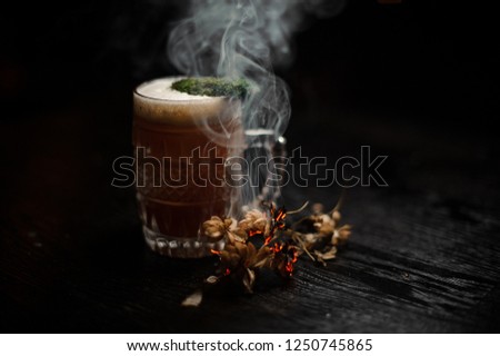 Beer glass with alcoholic drink decorated with moss and smoked dried flowers on the black table on the dark background