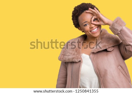 Young african american woman wearing winter coat over isolated background doing ok gesture with hand smiling, eye looking through fingers with happy face.