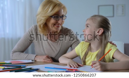 Smiling grandmother looking her granddaughter painting with colored pencils 