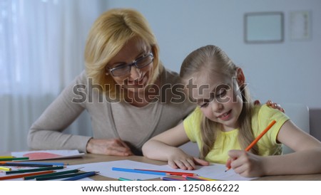 Caring retired woman looking at cute granddaughter painting with colored pencils