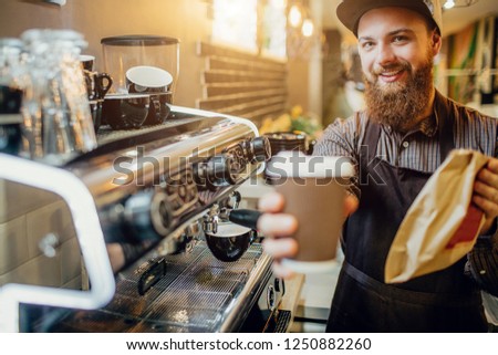 Picture of young bearded man holding cup of coffee and bag with food. He looks on camera. Guy stand in kitchen at coffee machine.