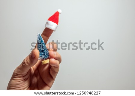 The man's hand holds a souvenir Christmas tree. Santa Claus hat on the index finger. The concept of a fun new year. Copy space. Gray background.