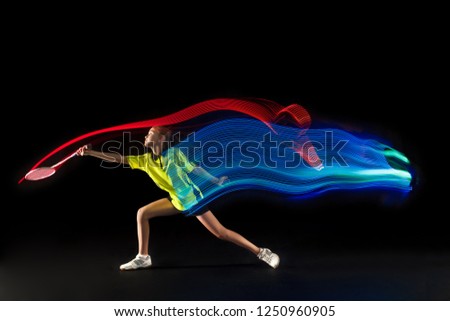 The one caucasian young teenager girl playing badminton at studio. The female teen player on black background in motion with flashes of  neon light