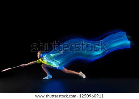 The one caucasian young teenager girl playing badminton at studio. The female teen player on black background in motion with flashes of neon  light