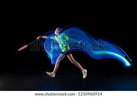 The one caucasian young teenager girl playing badminton at studio. The female teen player on black background in motion with flashes of neon  light