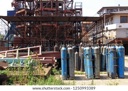 A photograph of a boat on land surrounded by rusty steel scaffolding with an array of blue gas tanks in the foreground of the photo.