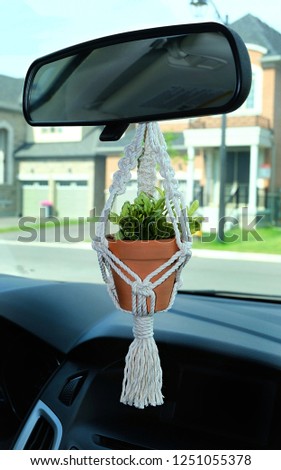 A hand-made mini macrame plant hanger made out of 100 percent cotton, holding a ceramic pot with a faux (fake) plant inside. The macrame is a car accessory decoration.