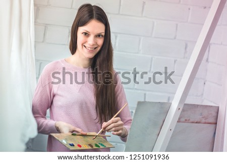 Beautiful young woman painting a picture on an easel. Holds a brush in her hands and palette of paints