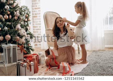 Happy young mother and her  little daughter in nice dress sit near the New Year's tree and open  New Year's gifts in the light cozy room